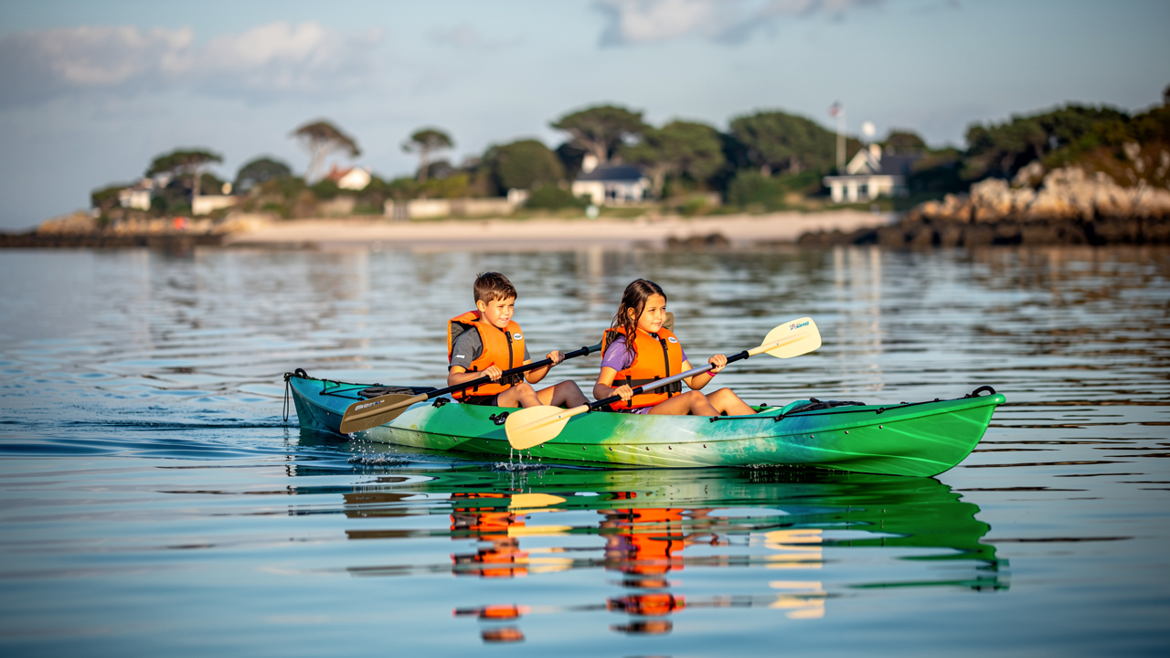 Un garçon et une fille portant des gilets de sauvetage pagaient dans un kayak biplace sur l’eau calme du Golfe du Morbihan, avec la presqu’île de Rhuys en arrière-plan.